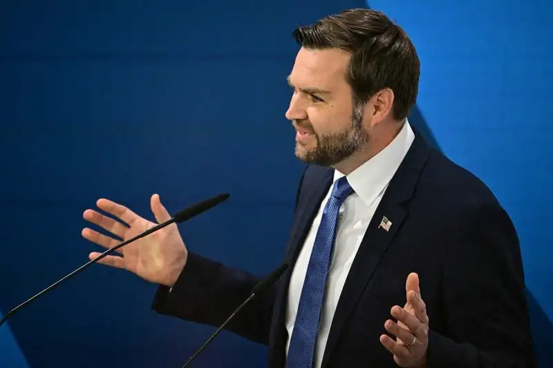 J.D. Vance gestures with both hands open as he speaks into a microphone during the Munich Security Conference. Vance wears a black suit with a blue tie.