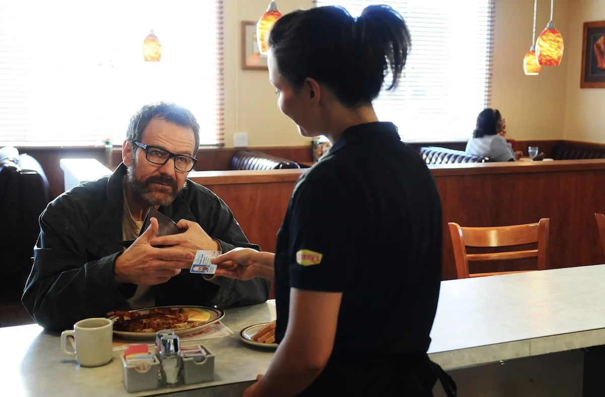 A man with dark hair and glasses sits at the bar in a diner and shows his identification to a waitress, a young woman with dark hair tied up. She is wearing a dark restaurant uniform with a Denny’s logo on the sleeve.