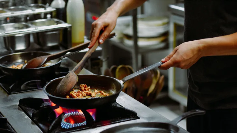 Hands stirring meat with wooden spoon in cast iron pan over gas range