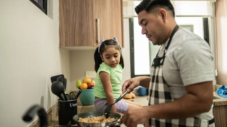 Man tends to a pot on stove while his young daughter sits on the counter next to him