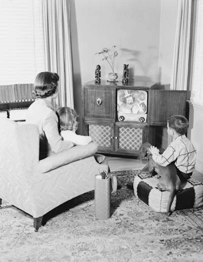 A black-and-white photograph of a mother and her two children watching television in the living room.