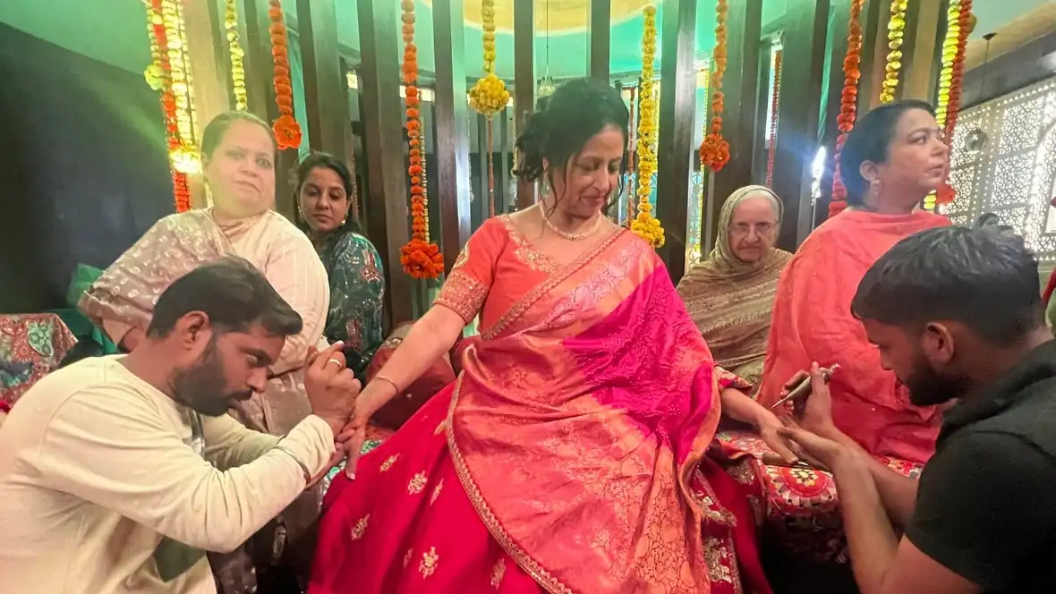 Sarab sits with her arms outstretched. She is having henna painted by assistants who are gathered around her. 