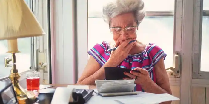 An elderly woman sits at a small table with documents. She is holding a checkbook and has a puzzled expression.