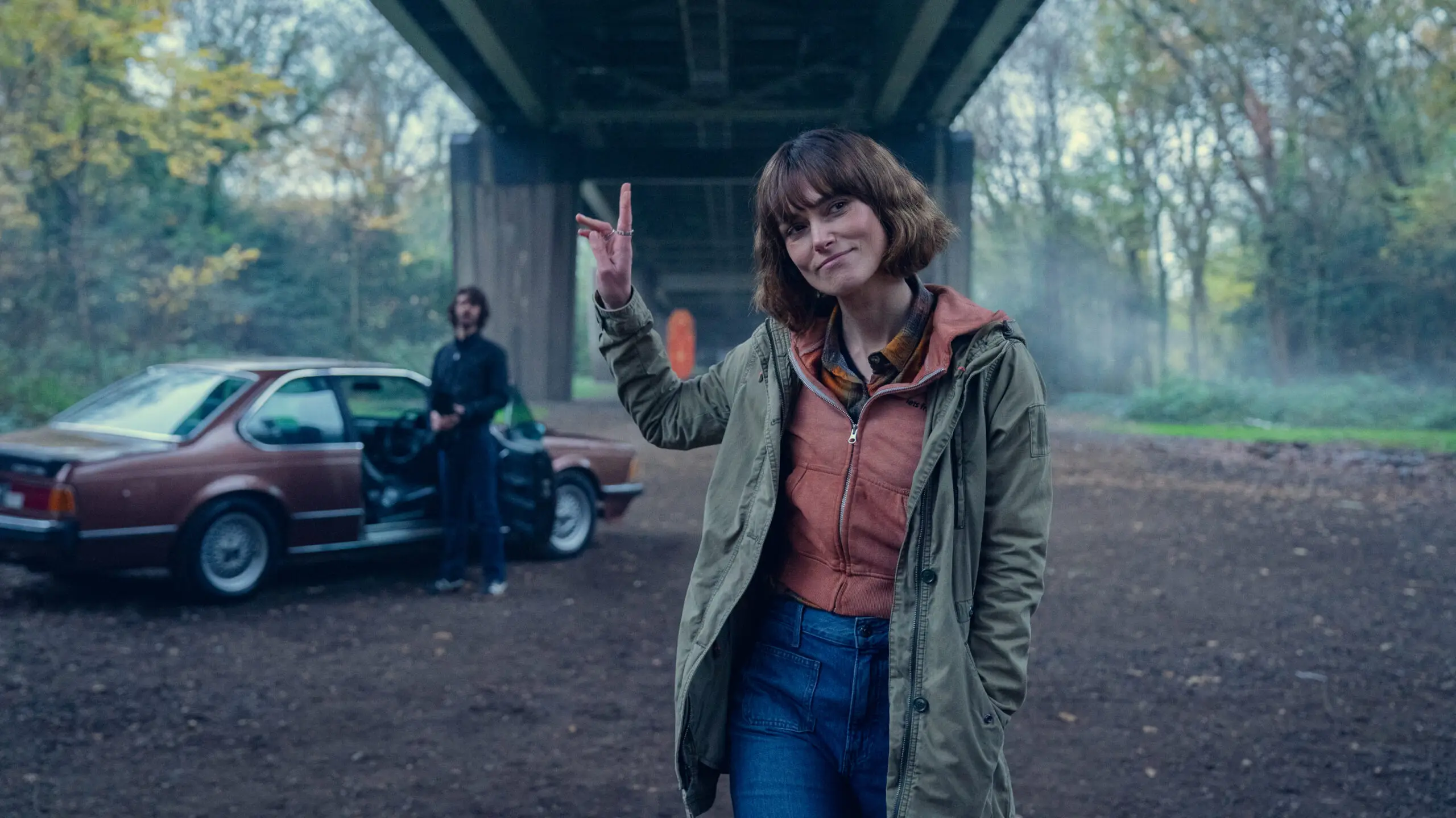 Woman giving peace sign near a car and man.