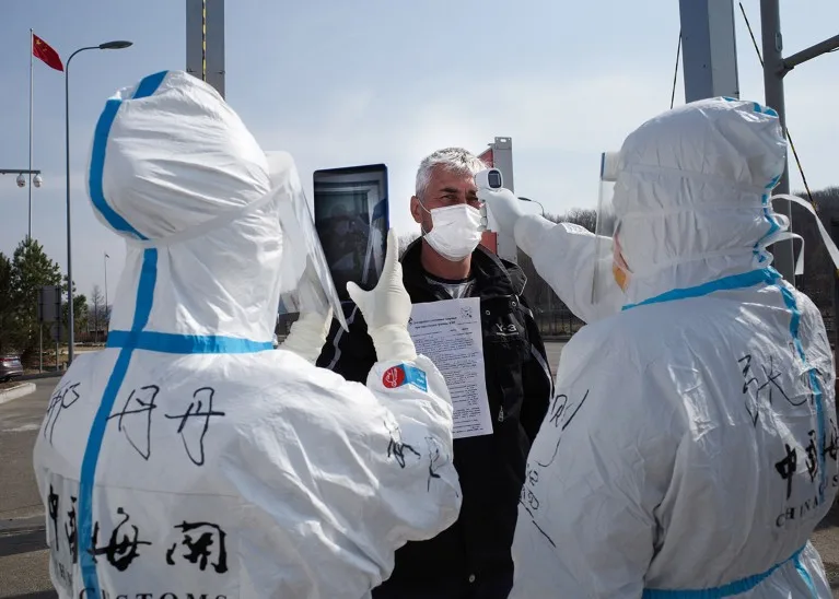 Two people wearing hazmat suits photograph and check the temperature of a man wearing a face mask.