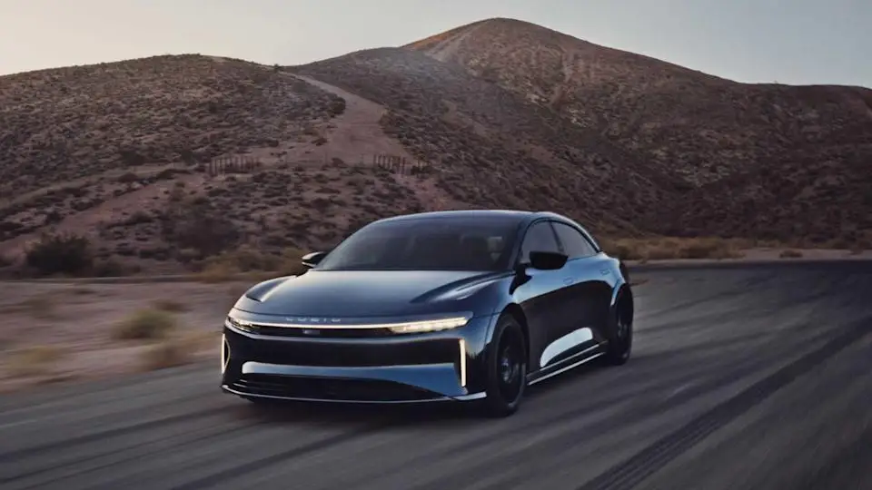 Full view of a blue Lucid Air Sapphire driving on a country road with a mountain in the background.