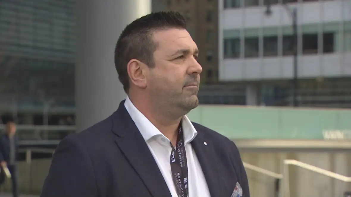 A white clean-shaven man speaks near a large building.