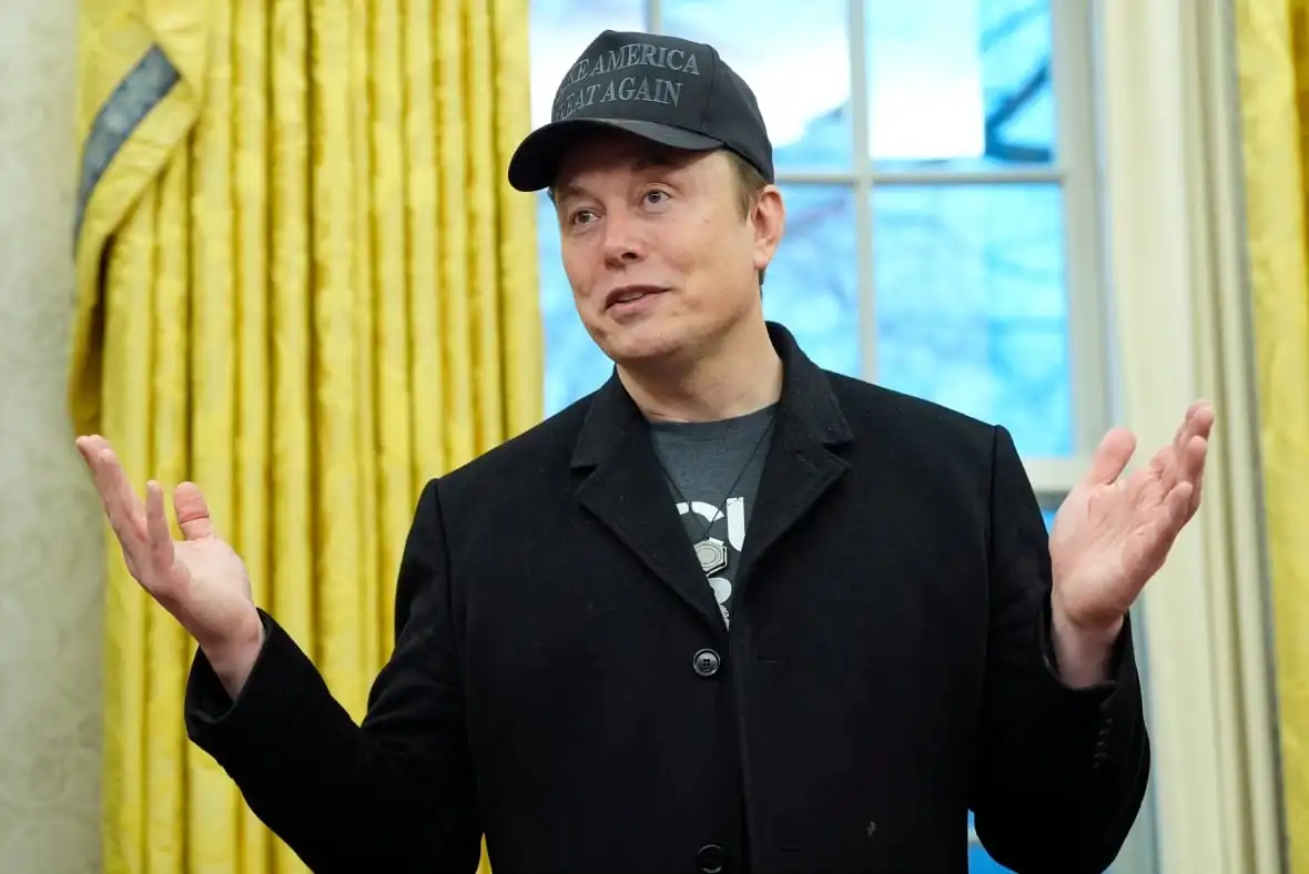 A clean-shaven man wearing a dark blazer, T-shirt and baseball cap extends his arms while speaking indoors.