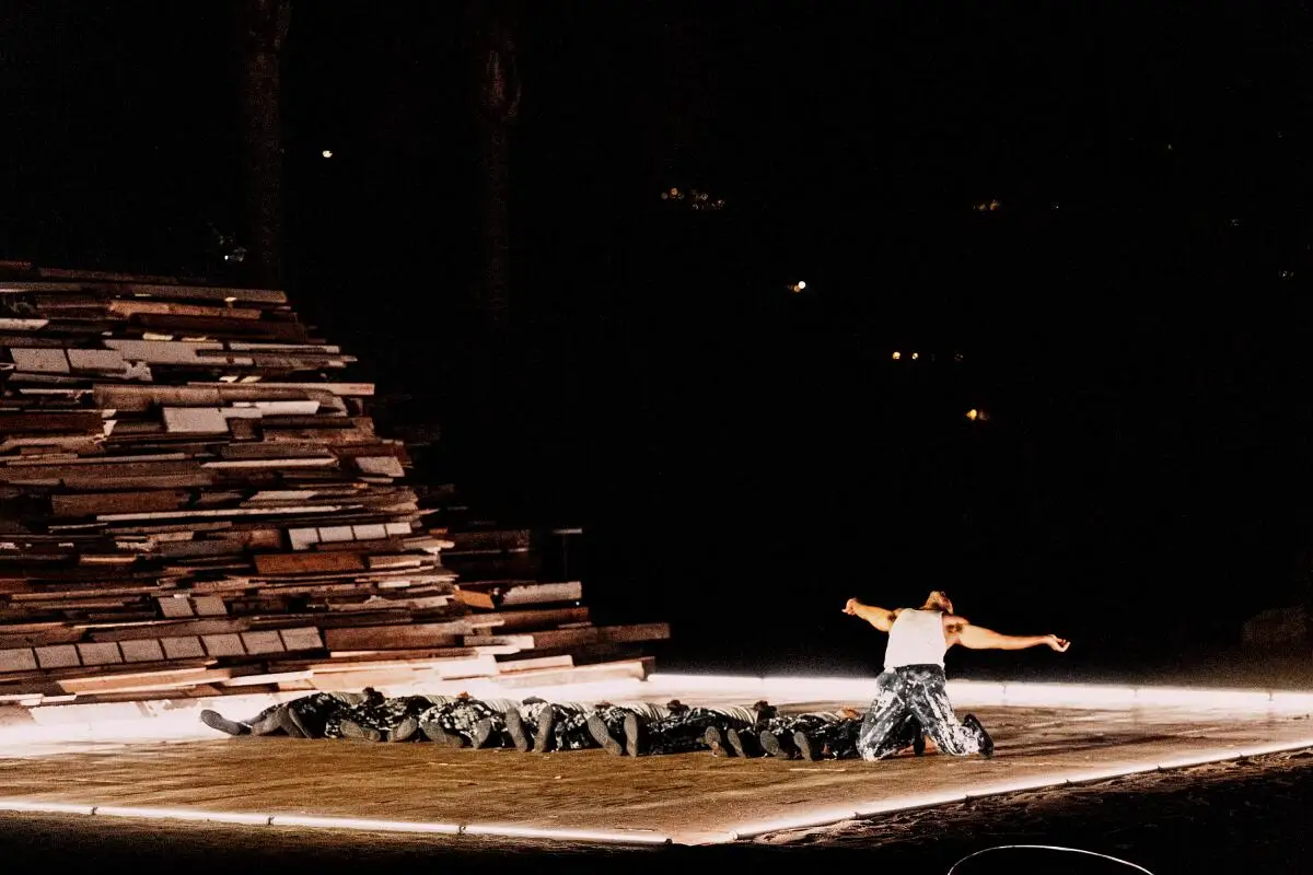 A man onstage on his knees leans back, arms extended, next to a pile of boards.