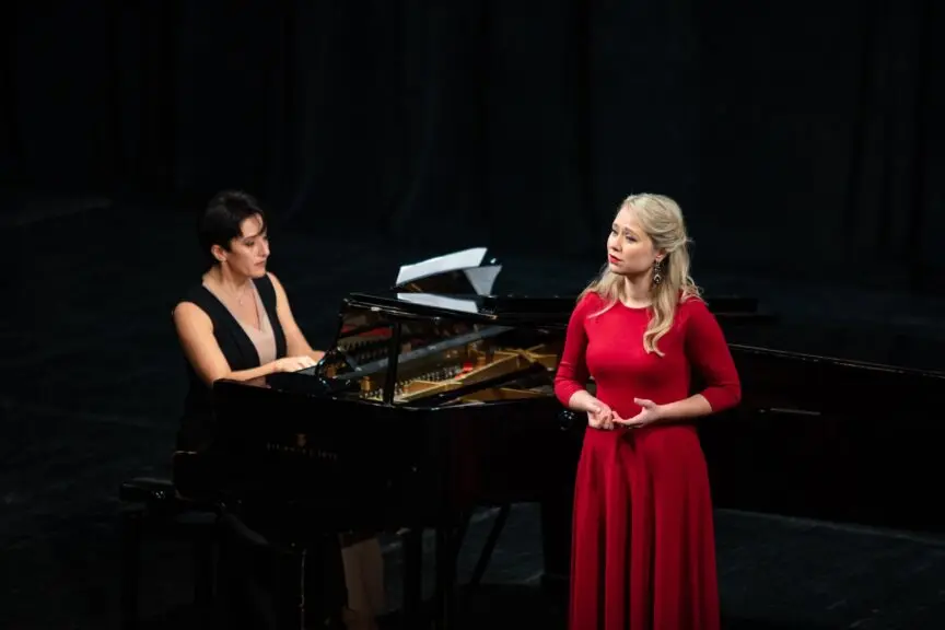 Viktoria, an opera singer, is performing on stage next to a seated male pianist. The background is dark, with the light focussed on the two people. Viktoria is a white woman with long, blonde hair, wearing a long red formal dress with long sleeves.