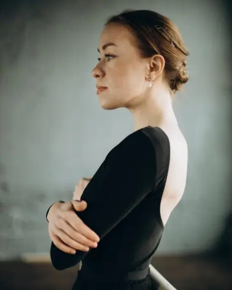 Nathalie is in a grey room pictured from the side with her arms folded, facing left.  She is a white woman with light brown hair in a low bun,  wearing a backless black dress with long sleeves.