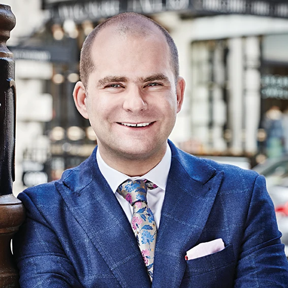 Man in a blue suit and floral tie smiles while standing in front of a blurred street background.