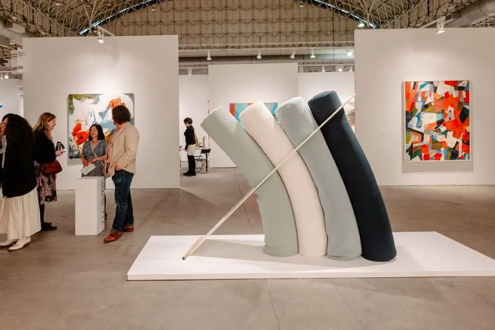 A sculptural artwork composed of five large, curved, soft cylindrical forms leaning together with a pole for support, surrounded by abstract paintings and fairgoers in conversation inside an EXPO Chicago exhibition booth.