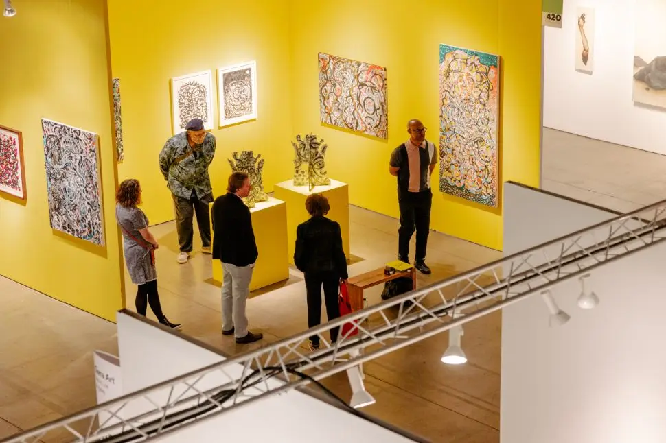 A brightly lit gallery booth with vivid yellow walls at EXPO Chicago, showcasing colorful abstract paintings and expressive sculptural works, as several visitors engage with the art and each other.