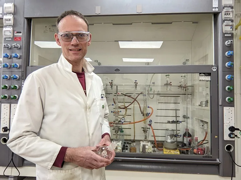 A man stands in a lab wearing a white jacket and goggles