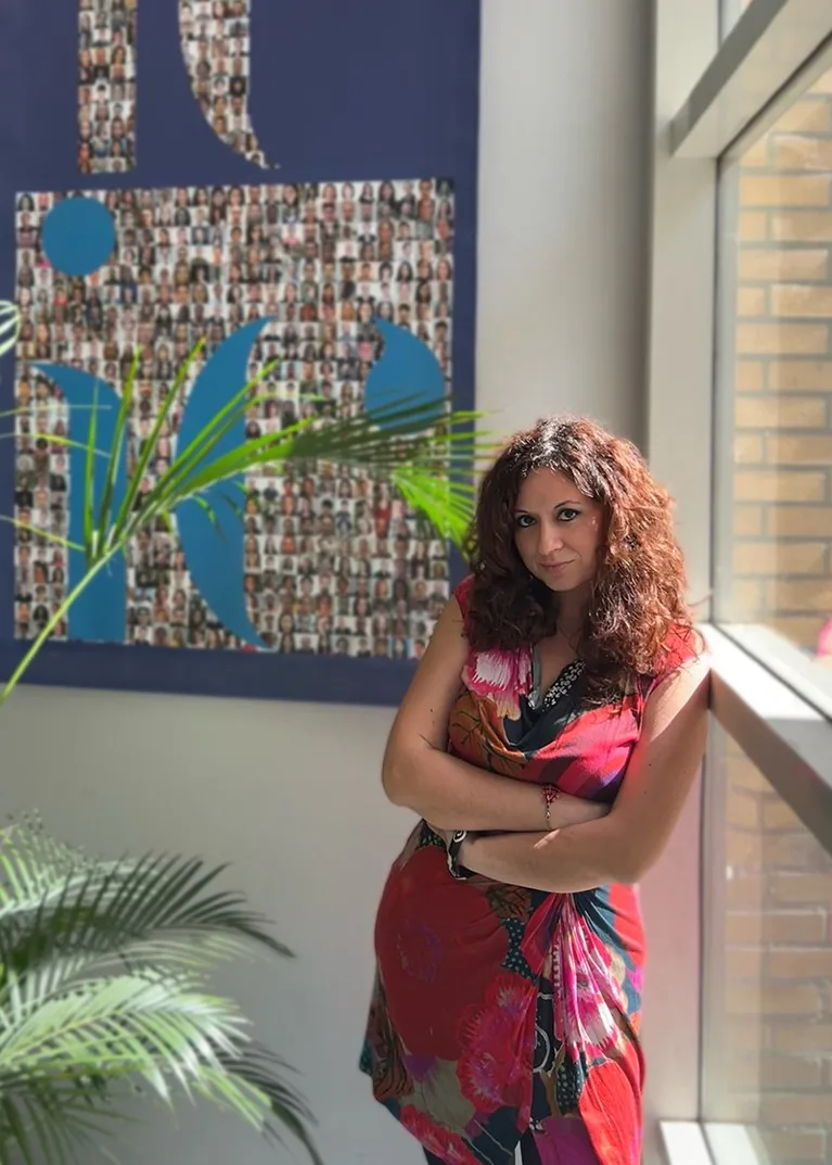 A woman stands by a window looking directly at the camera