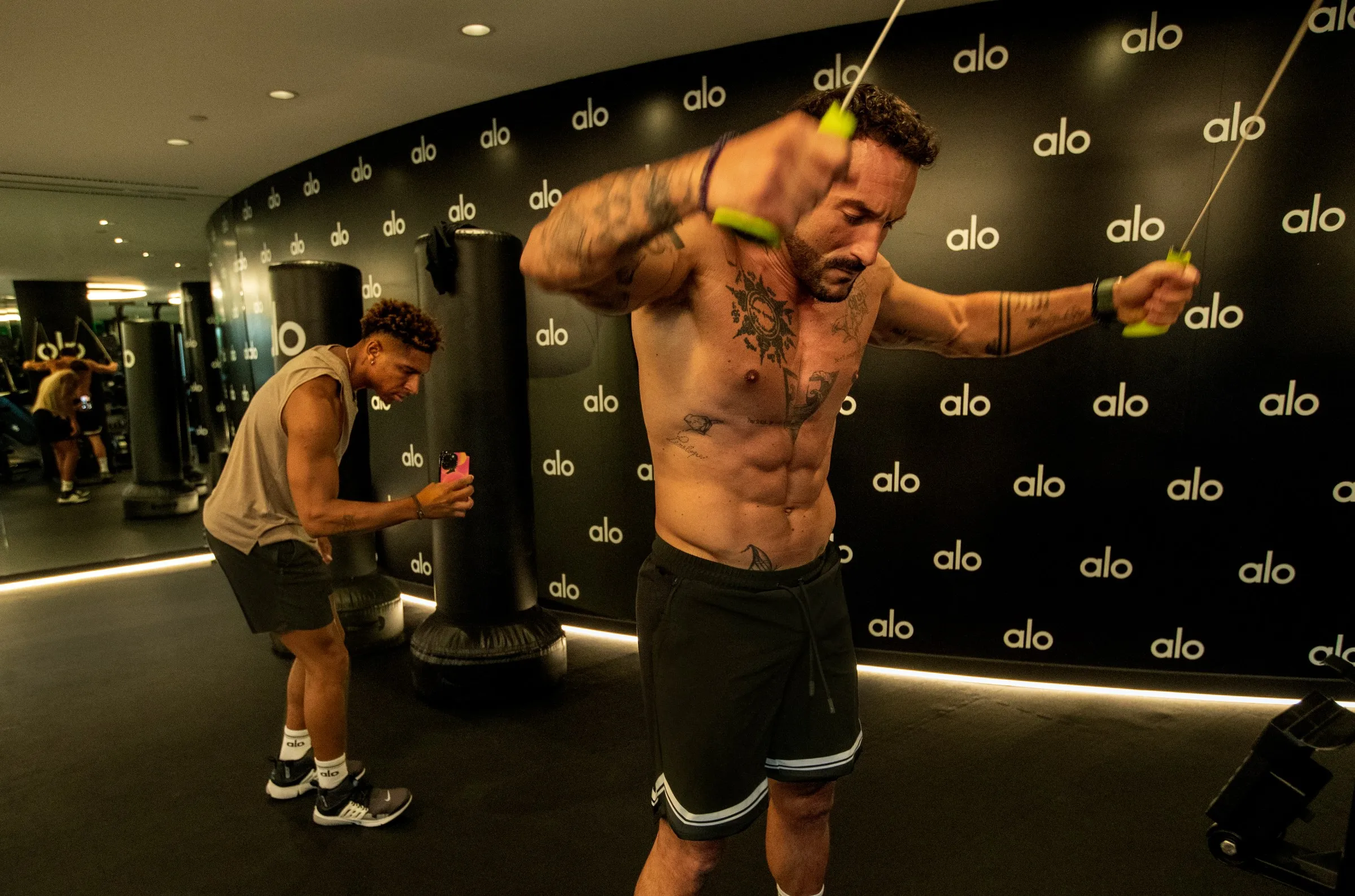 One muscled man exercising while another records him.