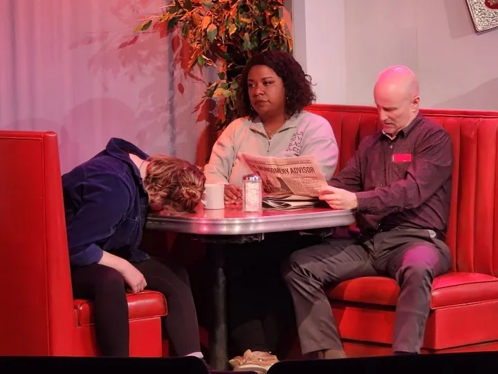 An actor rests their face on a diner table in frustration while two others look on.