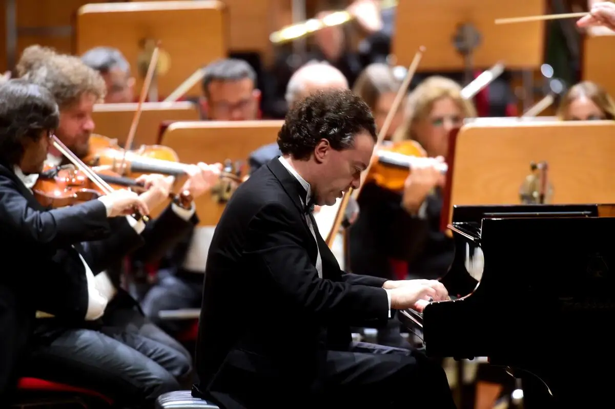 A man in formal attire play the piano with an orchestra.