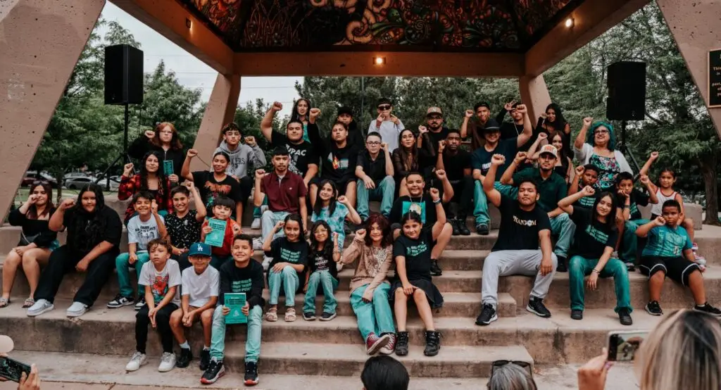 A large group of kids and teens pose, fist raised, under the plaza kiosko in Denver's La Raza Park