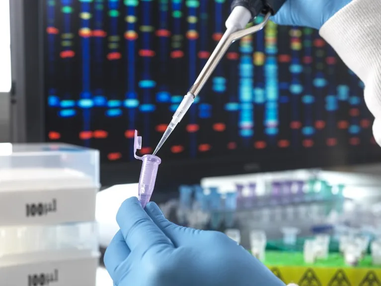 A researcher in PPE pours a DNA sample through a pipette into a test tube, with genetic results on screen in the background.