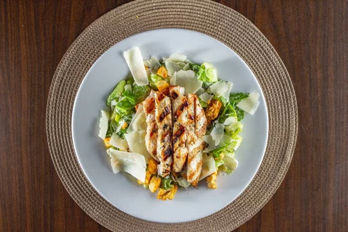 Grilled chicken breast on a Caesar salad with lettuce, croutons, and shaved Parmesan cheese on a round white plate