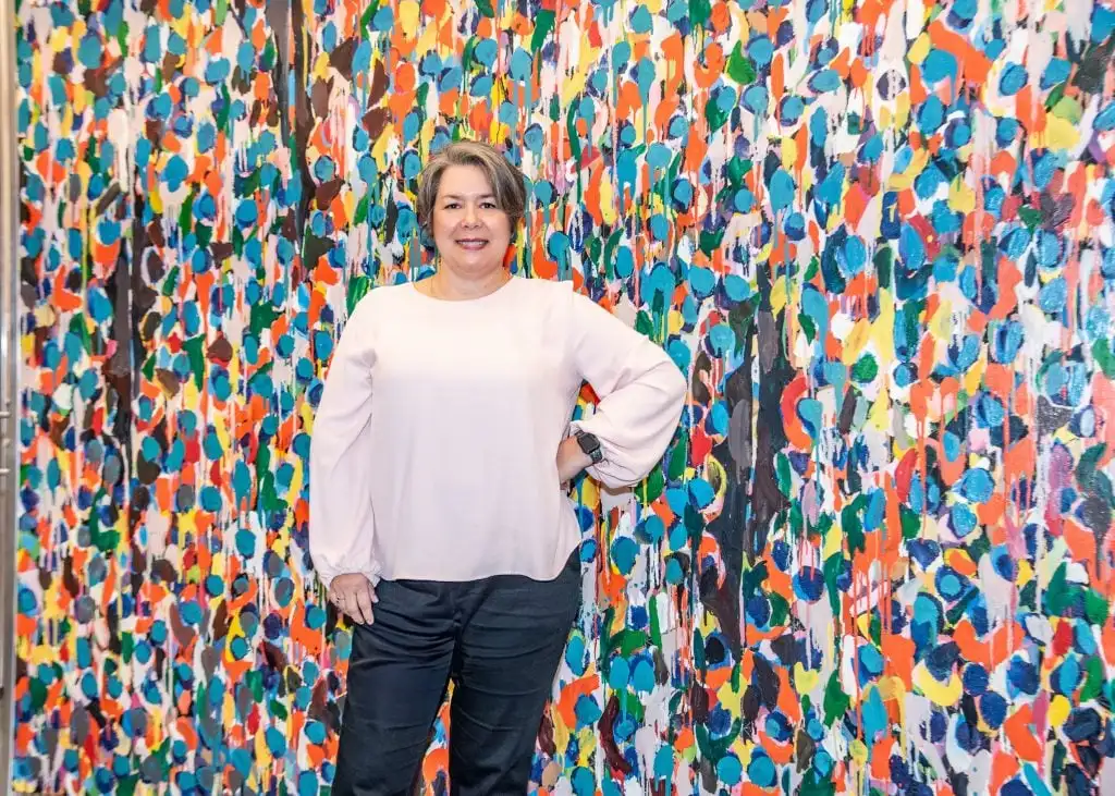 A Latina woman, Mónica Ramírez-Montagut, poses for the camera in front of colorful artwork