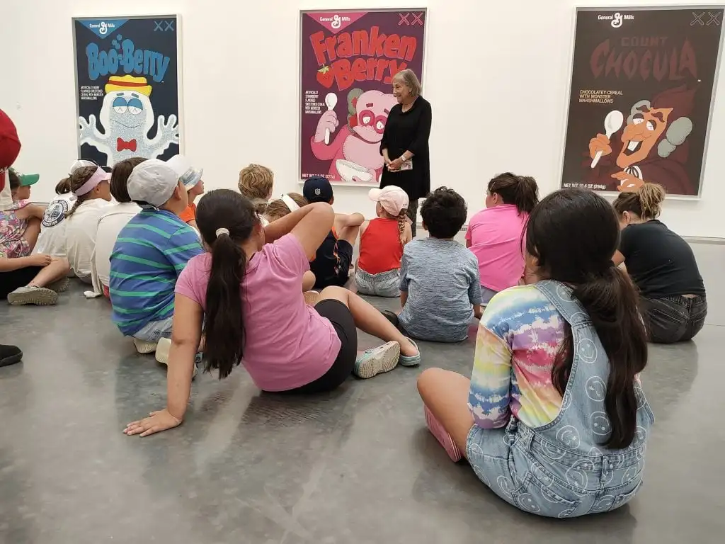 A guide stands in front of paintings on the wall and speaks to a seated group of young students