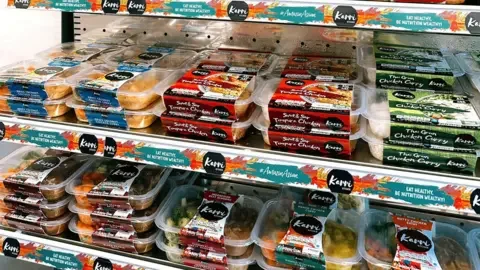 Karri Kitchen A range of ready meals across two shelves in a shop. 