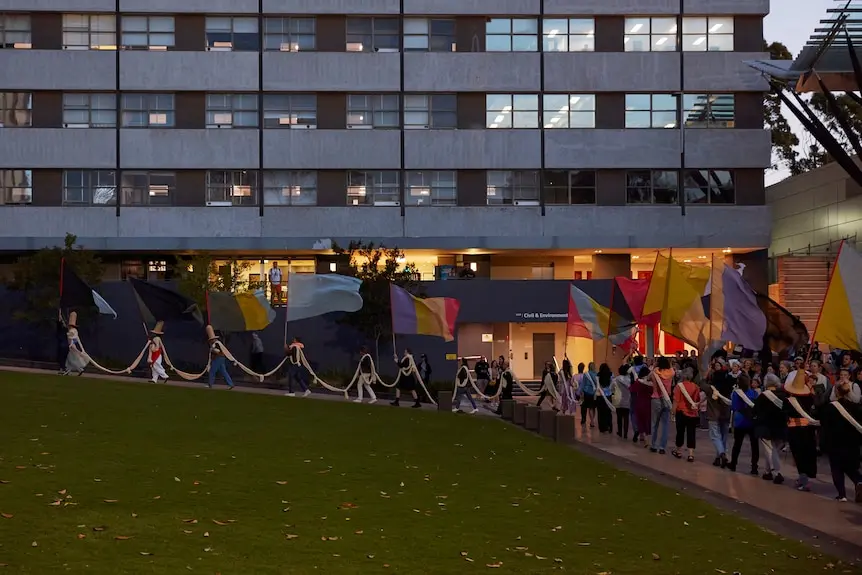Group of people all holding a large, long piece of cloth walk through green area beneath a building as it gets dark.