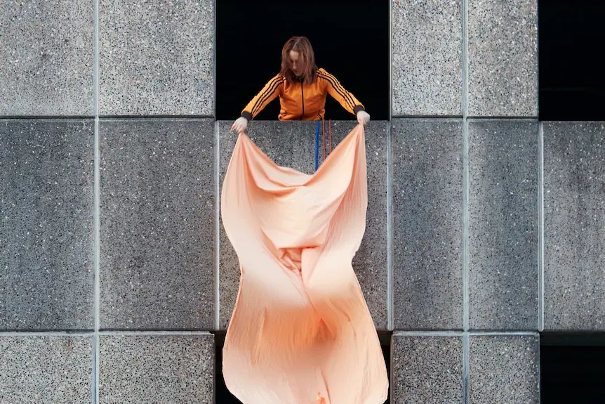 A woman hangs an orange piece of silk from a high open square in a cement building.