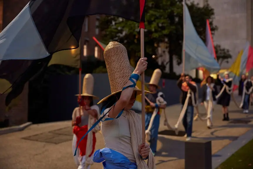 Person wearing straw hat holds large silk material on stick with blurred bodies walking behind her.