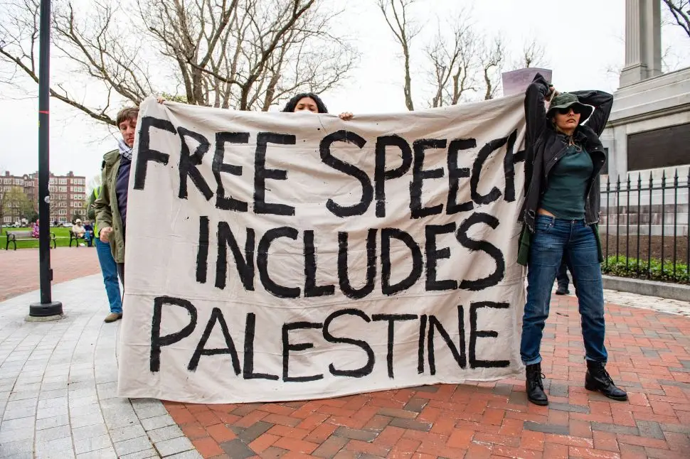 Three protesters hold a large banner reading “Free Speech Includes Palestine” during a demonstration in a city park near a monument.