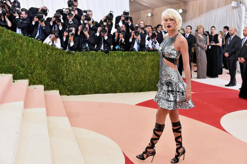 Taylor Swift in a metallic dress by a staircase near dozens of photographers wearing black.