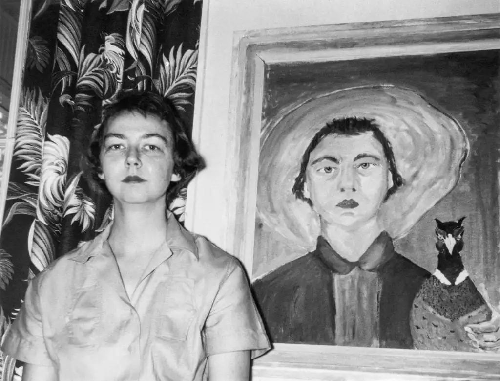 O’Connor stands beside self-portrait painting holding bird, both wearing solemn expressions and dark clothing.