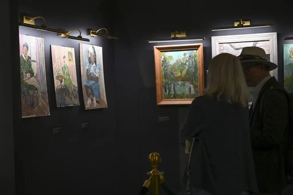 Two people observe Flannery O’Connor’s expressive portraits and landscapes illuminated in a dimly lit gallery.