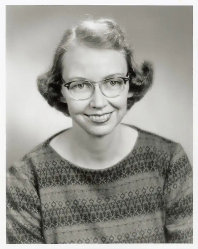 Flannery O'Connor with glasses and patterned sweater smiles in formal, vintage black-and-white studio portrait.