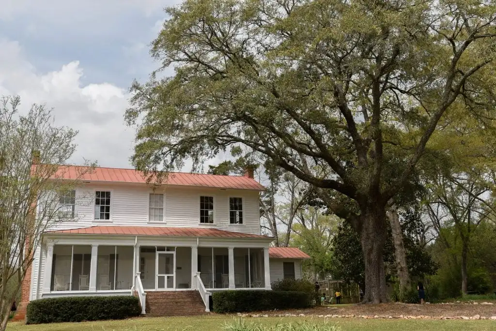 Historic white farmhouse with red roof and large oak tree, surrounded by green lawn and trees.