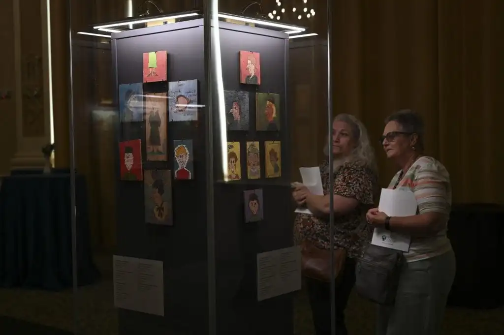 Two women observe glass display case of small colorful portrait paintings mounted in museum exhibition.