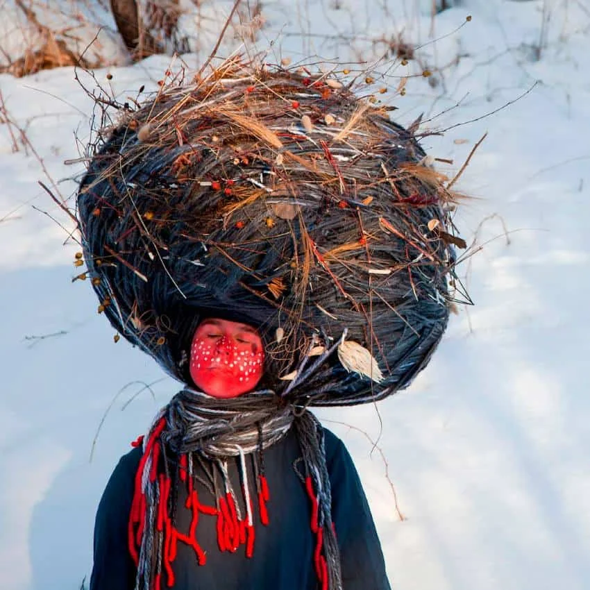 Photoshopped image of a woman with her face painted red with white dots of paint across her cheeks and nose. She is wearing a headdress made of yarn, straw and other natural matter and a cloak. Her photo is superimposed on a woodsy area with thick snow.