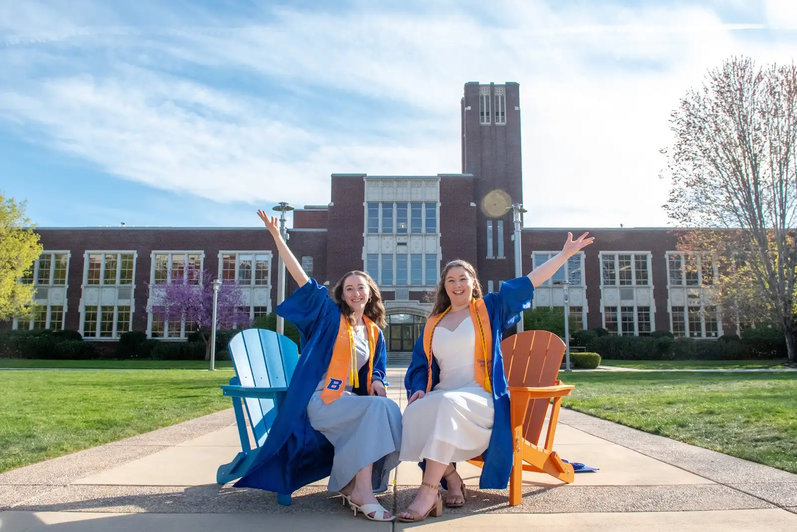 Two women in commencement regalia pose with arms up outside Administration building