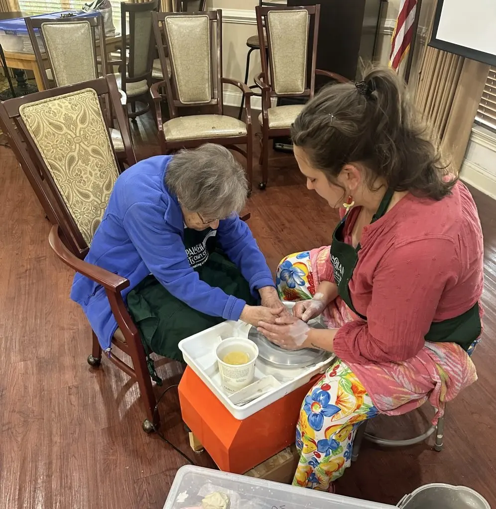 An elderly woman works on creating an art piece with the help of a younger woman