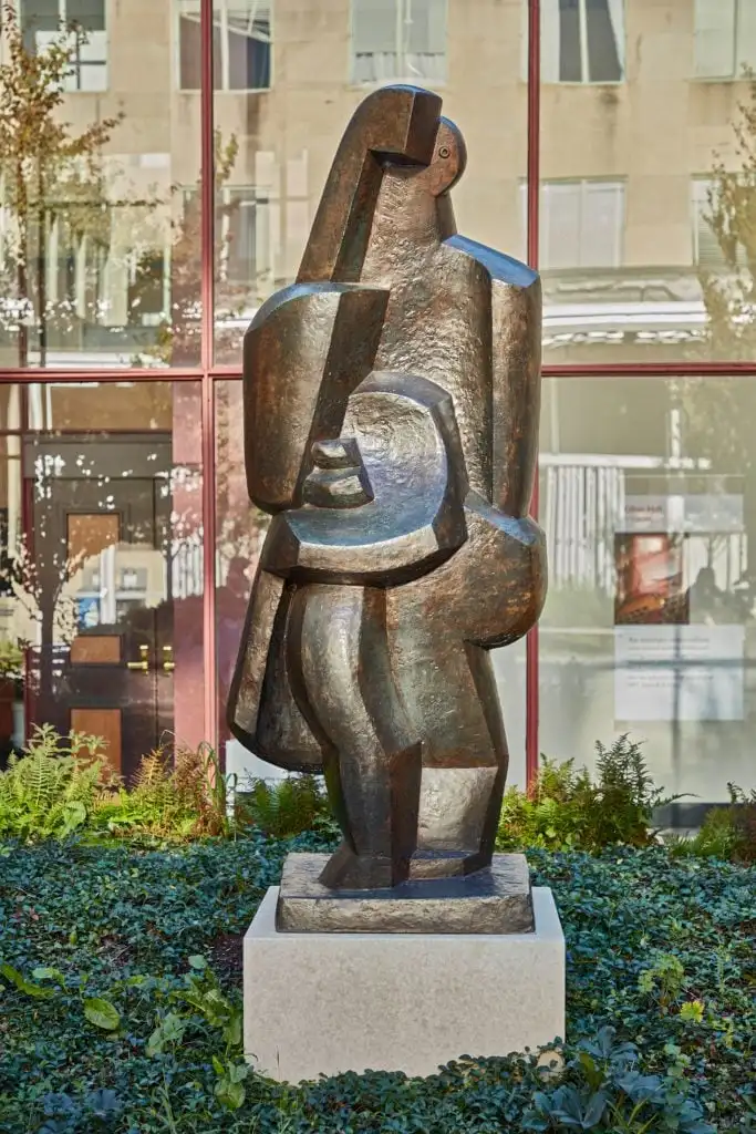 Cubist-style bronze sculpture of woman and child stands outside glass-walled building with plantings below.