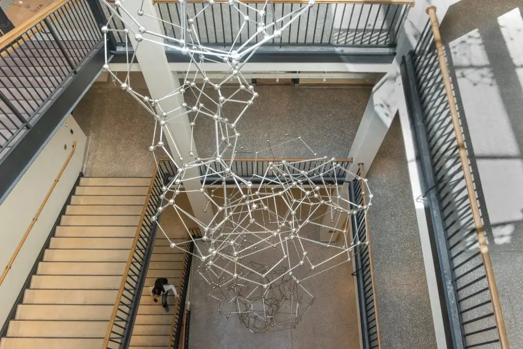 Intricate geometric sculpture hangs through multi-level stairwell inside building, viewed from above.
