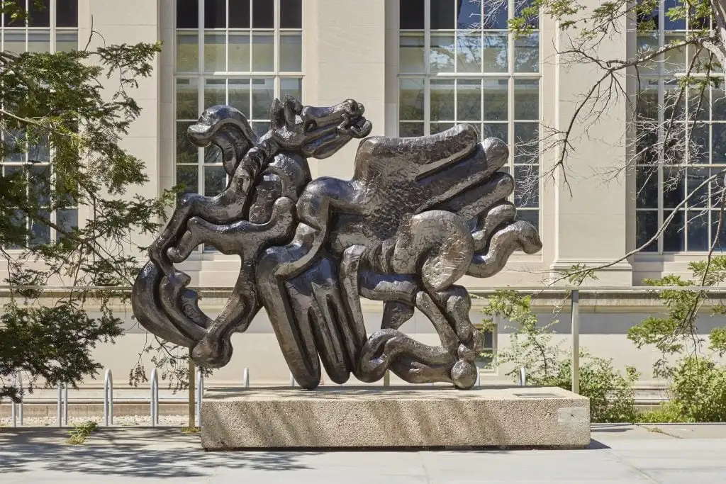 Shiny metallic sculpture depicts mythological horse and figures in dynamic pose outside neoclassical building.