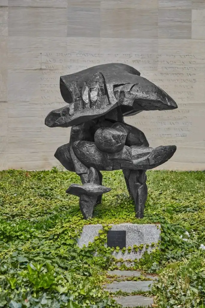 Abstract bronze sculpture with organic shapes stands in ivy-covered garden before engraved stone wall.