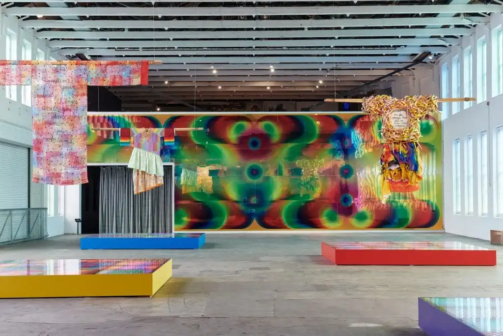 Colorful textile sculptures hang in a gallery with a vibrant, psychedelic mural and mirrored platforms.