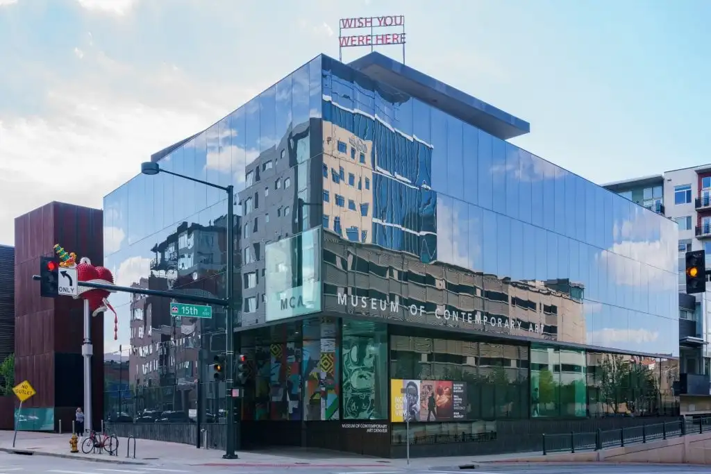 Glass-walled museum reflects cityscape, features rooftop “WISH YOU WERE HERE” sign and heart-shaped traffic sculpture.
