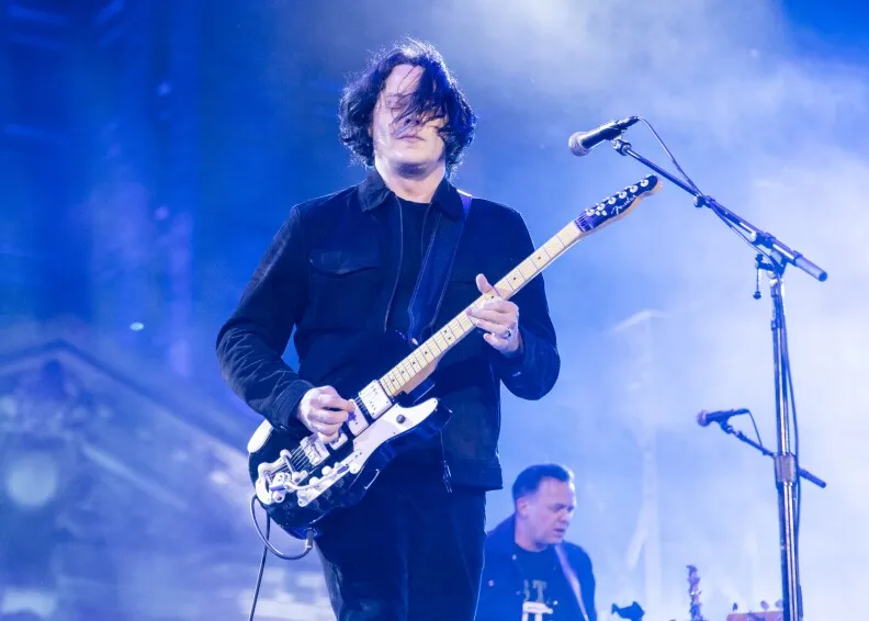 Jack White plays a black and white electric guitar on stage during a performance. He's wearing all black, and the stage lights are shades of blue. Behind him, another man is performing in the band too.