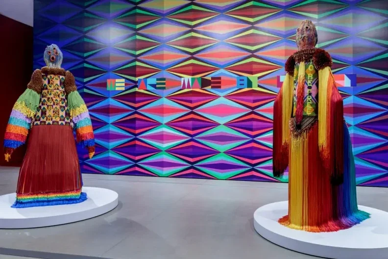 Two multicolored, human-like statues depicting ancestral figures in front of a bright, colorful patterned wall in a museum.